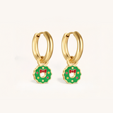 Gold hoop earrings with green wreath charms featuring red bows on a light pink background