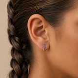 A close-up side profile of a woman's ear wearing a small silver-toned earring with a pink gemstone and a decorative bow detail, against a warm beige background.
