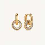 A pair of gold huggie hoop earrings with a smaller concentric ring partially paved with clear stones, shown on a light cream background.