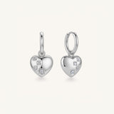 A pair of silver heart-shaped hoop earrings with small clear gemstones, shown on a light cream background.