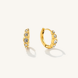A pair of gold huggie hoop earrings with dangling star-shaped charms embellished with clear crystals, shown on a light cream background.