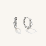 A pair of silver huggie hoop earrings adorned with clear round gemstones, displayed on a clean off-white background.